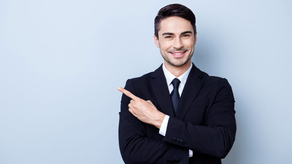 Close up portrait of young successful brunete stock-market broker guy on the pure light blue background, he is smiling, wearing suit with tie and is pointing on a copyspace with his finger
